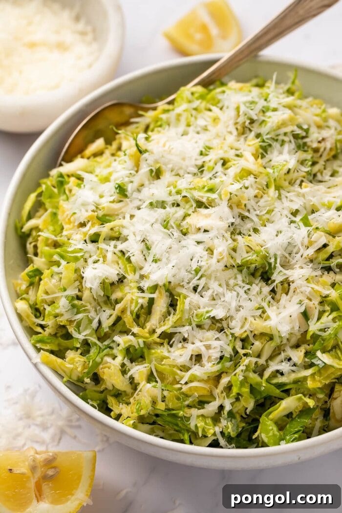 Angled side view of a bowl of shaved Brussels sprouts salad with lemon and parmesan, garnished with lemon wedges and extra cheese