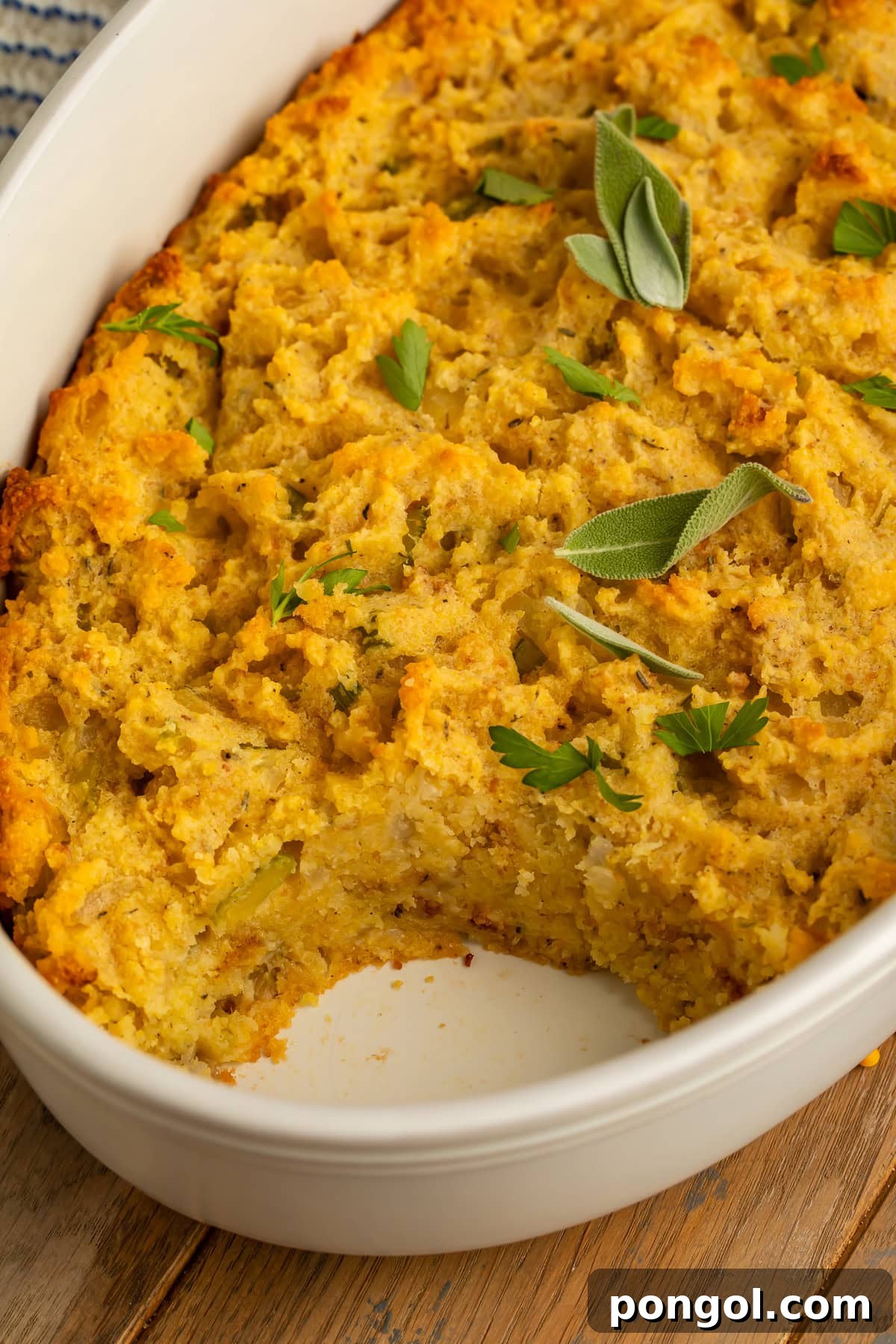 Golden cornbread stuffing in a white oval baking dish with a portion removed, garnished with fresh sage and parsley.