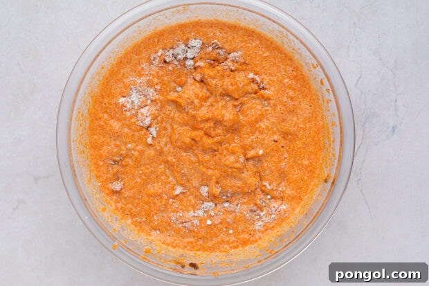 Dry ingredients like flour and cinnamon being added to the carrot soufflé mixture in a mixing bowl.