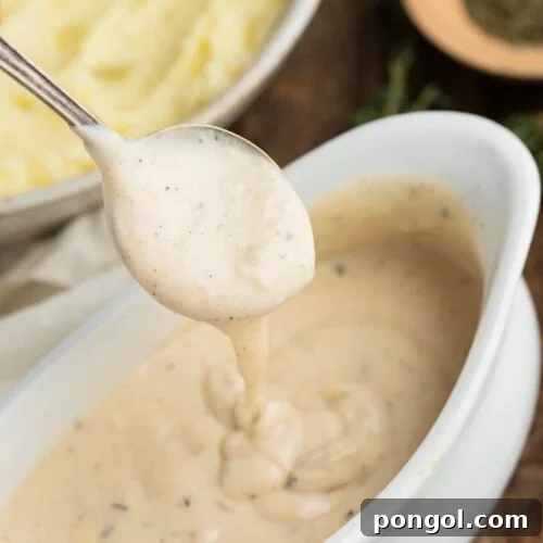 A spoonful of rich white gravy held above a gravy boat, with a large bowl of creamy mashed potatoes in the background.