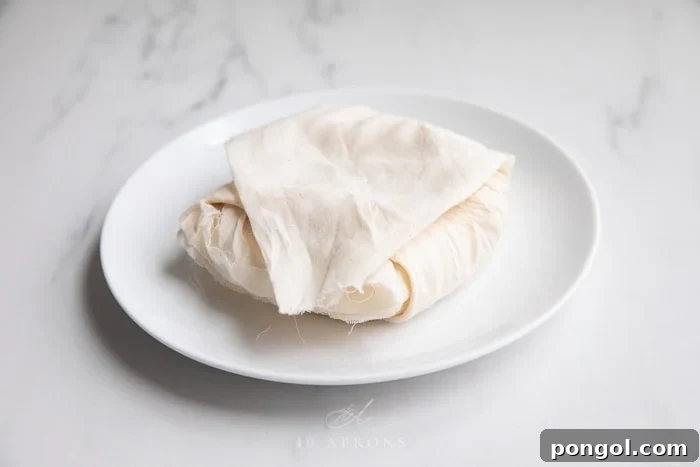 A tightly wrapped disc of paneer in cheesecloth, pressed under a plate weighted with two cans. This setup illustrates the pressing stage, where excess moisture is removed to firm up the homemade Indian cheese, crucial for its texture.