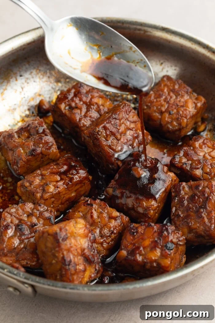 Tempeh cubes being coated with a savory marinade in a bowl, showing rich color and texture.
