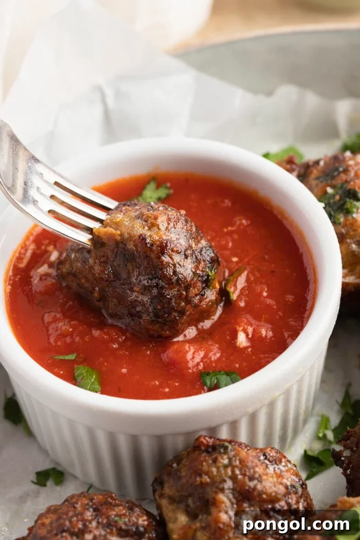 A single air fryer meatball on the end of a fork, being dipped into a small ramekin of rich red marinara sauce, highlighting its crispy exterior.