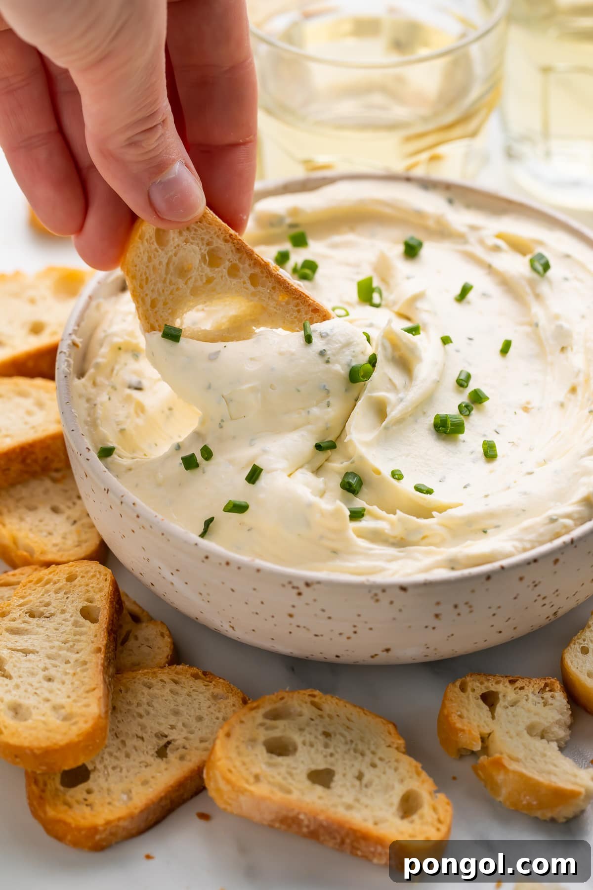 Herbed Garlic Cheese Spread from Scratch 3 A white woman's hand dipping a piece of crostini into a bowl of creamy homemade boursin cheese, highlighting its perfect texture for dipping.
