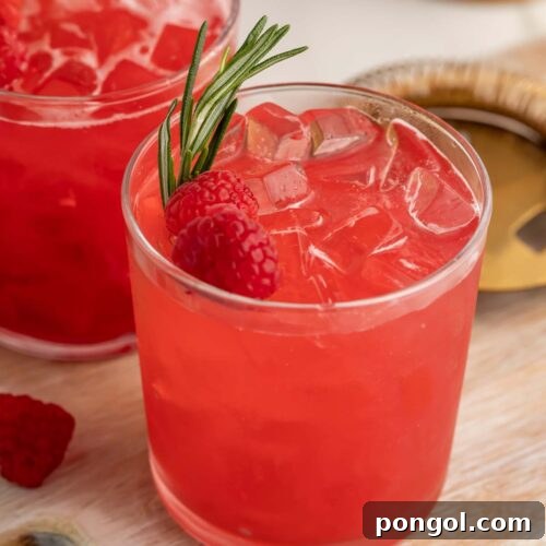 A bright red raspberry cocktail in a glass, garnished with a raspberry and sprig of rosemary.