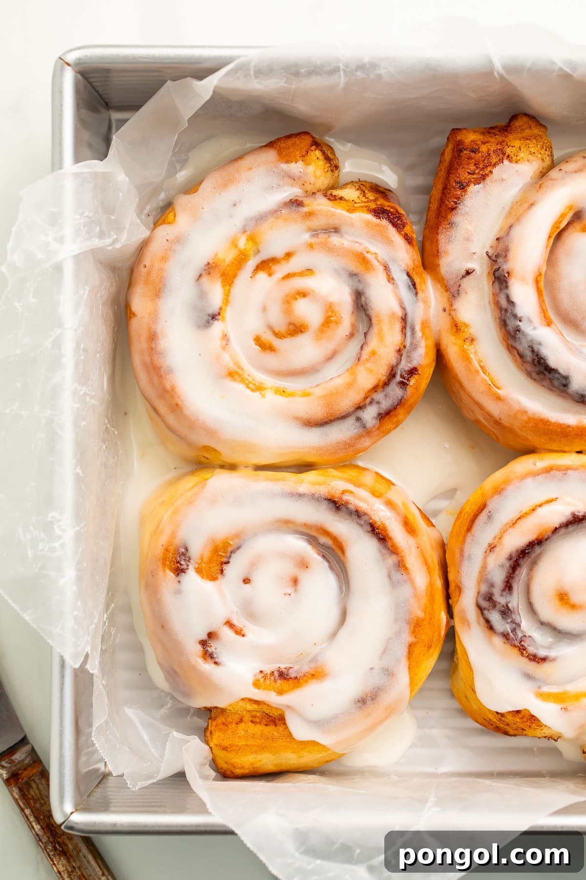 Four air fryer cinnamon rolls, generously topped with melty cinnamon roll icing, rest in a pan lined with parchment paper, inviting a delicious bite.