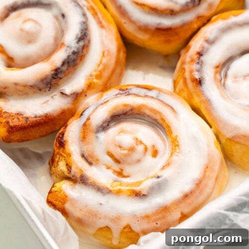 Four air fryer cinnamon rolls topped with melty cinnamon roll icing resting in a pan lined with parchment paper.