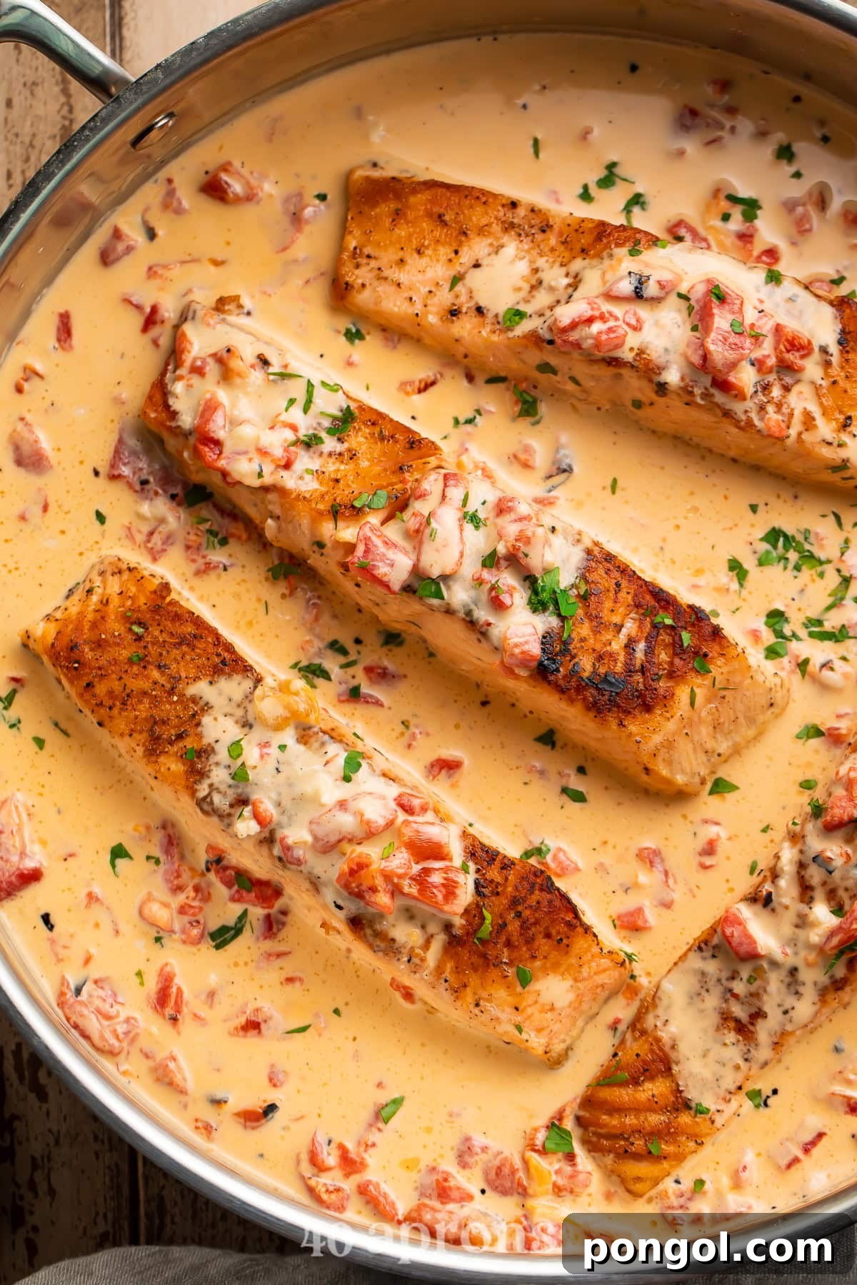 Four pan-seared salmon fillets in a large silver skillet with creamy roasted red pepper parmesan sauce and fresh parsley garnish.