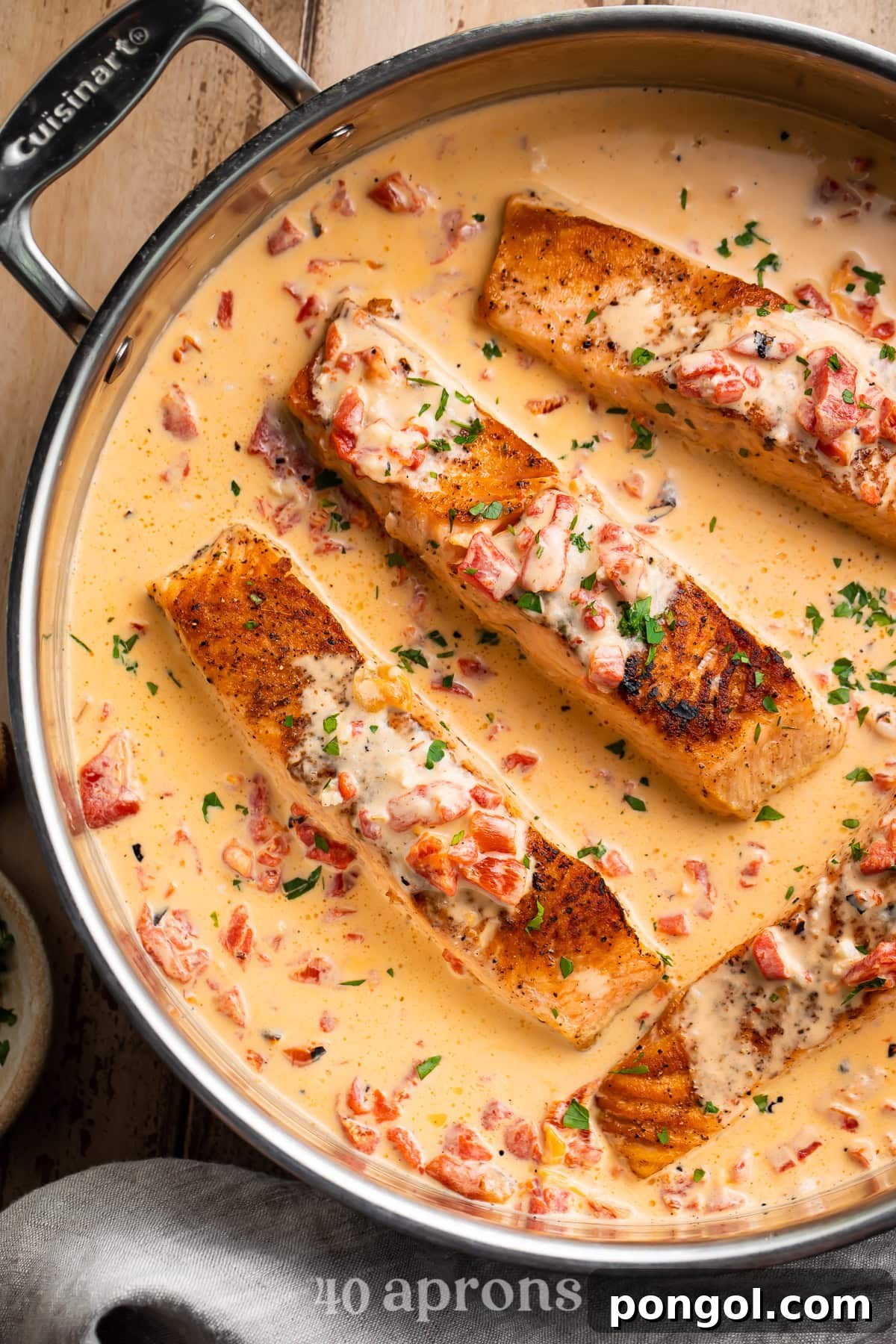 Overhead view of seasoned salmon fillets in a Cuisinart skillet covered in creamy red pepper sauce with herbs.