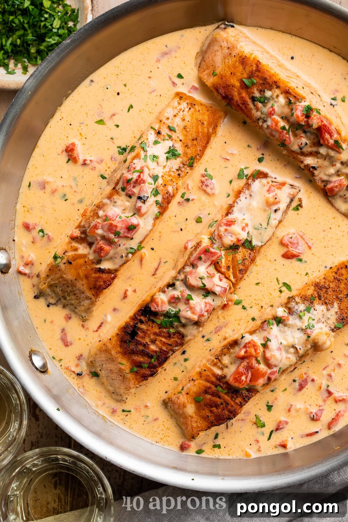 Sliced pan-seared salmon fillets in creamy red pepper parmesan sauce with fresh herbs and white wine glass.