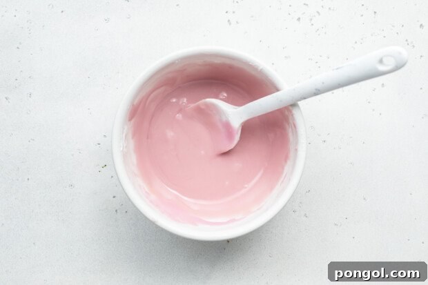 Overhead view of pink vanilla icing for chocolate gluten-free donuts in a large glass mixing bowl with a spoon.