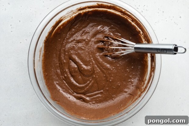 Overhead view of chocolate gluten-free donut batter in large glass mixing bowl with metal whisk.
