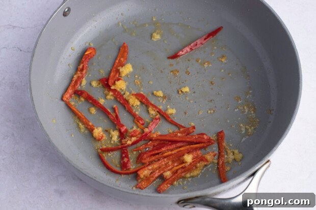 Red chilis, minced garlic, and grated ginger in large grey skillet on white background.