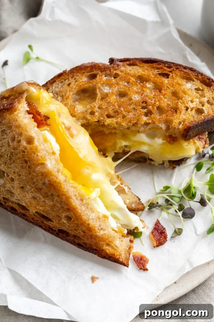 The Ultimate Breakfast Stack 3 A fried egg sandwich, sliced in half diagonally. One half rests on the plate while the other half stands upright to beautifully display the golden egg yolk and melted cheese.