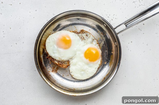 The Ultimate Breakfast Stack 6 Two fried eggs cooking in a large silver skillet with bacon fat, whites setting and yolks glistening.