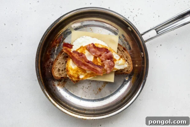 The Ultimate Breakfast Stack 7 Partially assembled fried egg sandwich in a large skillet, showing layers of bread, cheese, eggs, and bacon.