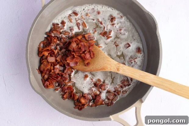 Overhead view of chopped bacon pieces in large grey skillet with wooden spoon and rendered bacon fat.
