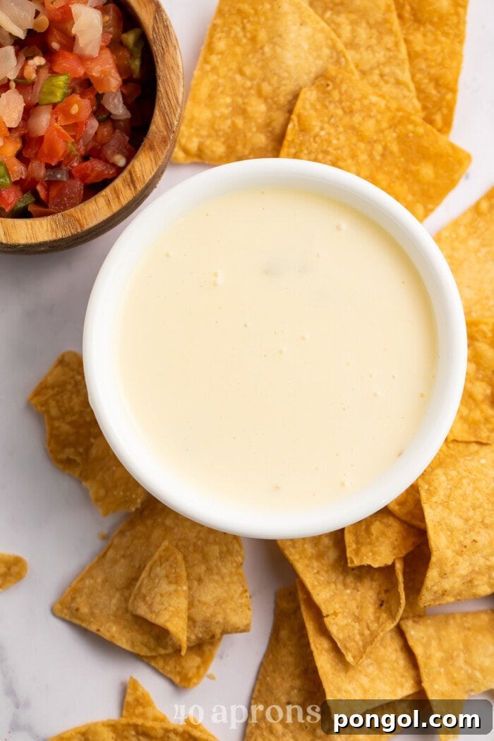 Velvety White Queso Dip 2 Overhead view of a bowl of white queso with a dollop of diced tomatoes in the center, surrounded by tortilla chips.