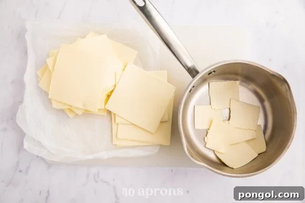 Velvety White Queso Dip 6 Overhead view of white American cheese on a cutting board next to a saucepan.