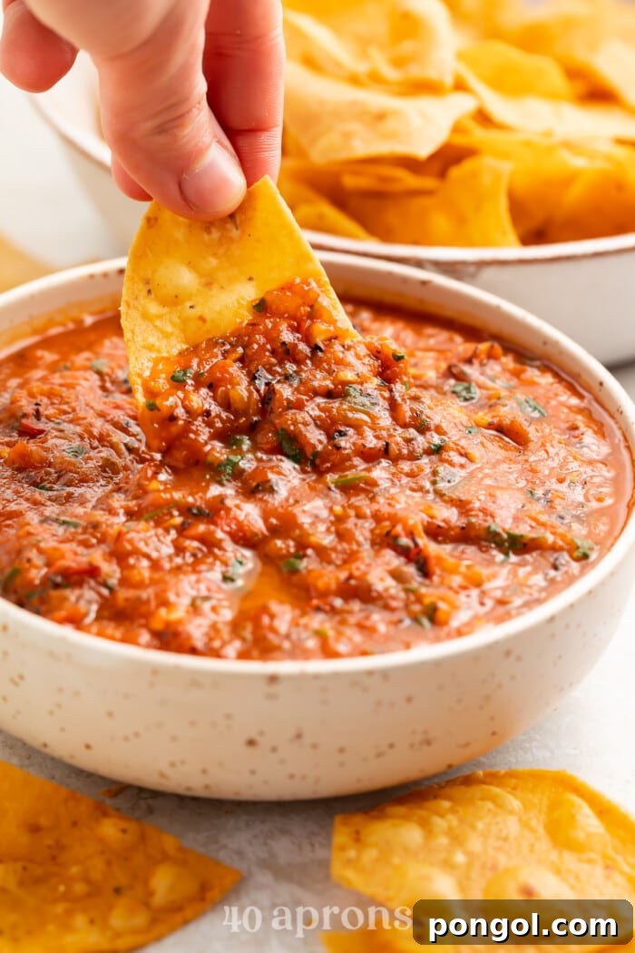 A tortilla chip being dipped into a white bowl containing red roasted salsa.