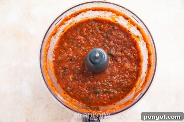 Overhead view of red roasted salsa in a food processor bowl on a light neutral countertop.