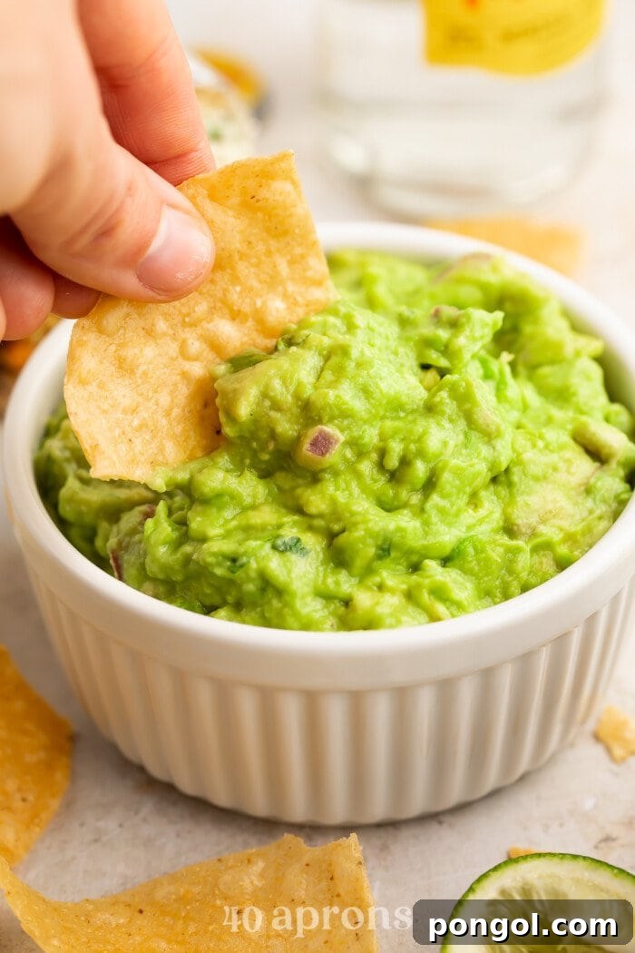 A woman's hand dipping the corner of a tortilla chip into a ramekin of homemade Chipotle guacamole, garnished with a sprinkle of red onion and jalapeño.