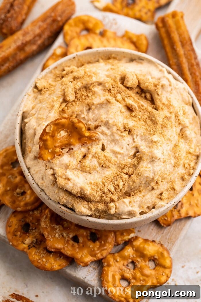 Angled view of a small white bowl of churro dip topped with crushed cinnamon cereal, surrounded by a variety of dippers like pretzels and fruit slices.
