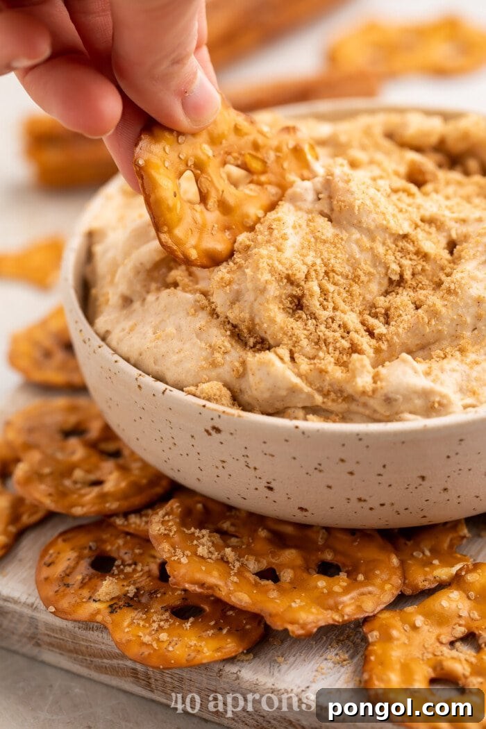 Side view of a hand scooping churro dip out of a small white bowl with a pretzel chip, highlighting the dip's creamy texture.
