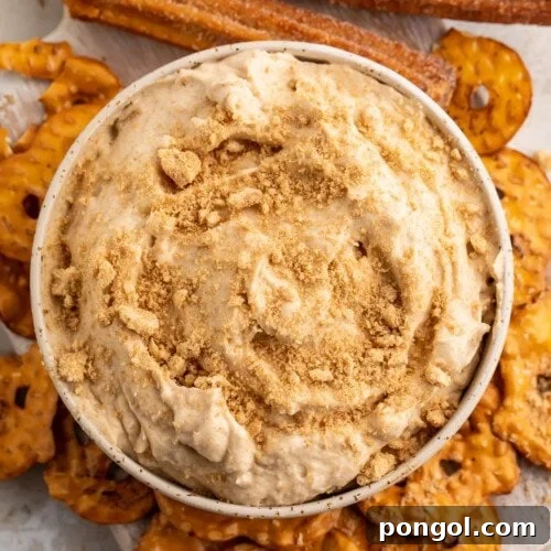 Overhead view of a small white bowl of churro dip surrounded by pretzel chips on a white countertop, highlighting its deliciousness.