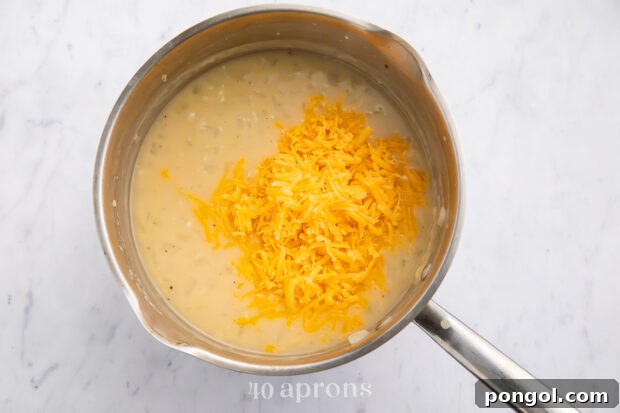 Overhead view of chicken broth, garlic, onions, flour, and shredded cheddar cheese in a medium silver saucepan with a silver handle, forming the cheese sauce.