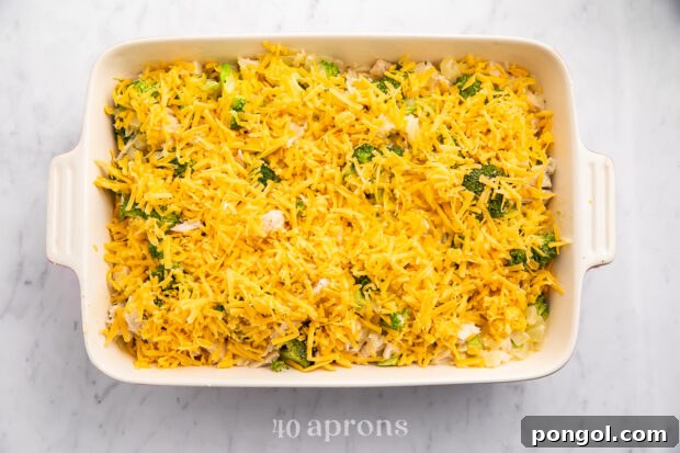 Overhead view of cheesy chicken, broccoli, rice casserole in a large white rectangular casserole dish, topped with cheese, before baking.