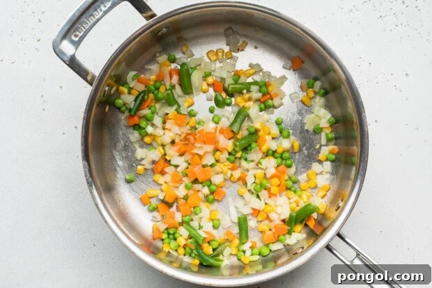 Overhead view of a large skillet with avocado oil, diced onion, and frozen mixed vegetables sizzling.