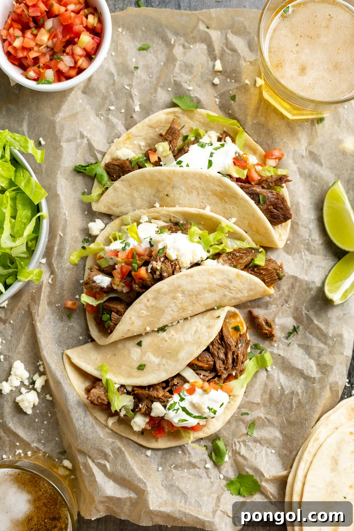 Overhead view of shredded beef tacos laying on a sheet of parchment paper, surrounded by toppings.