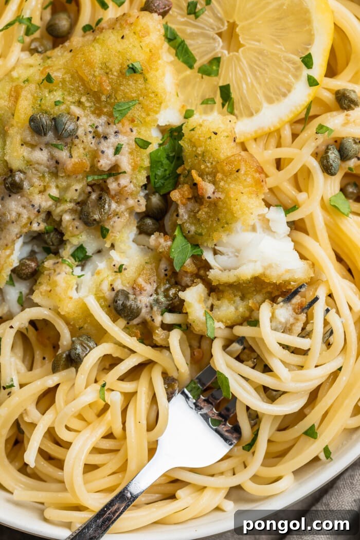 Close-up view of potato-crusted cod piccata with spaghetti noodles, lemon slices, capers, and fresh parsley.
