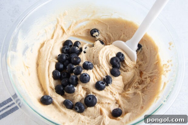 Fresh blueberries in gluten free blueberry muffin batter in large glass mixing bowl with spatula.