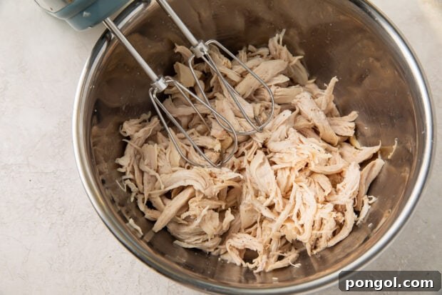 Shredded chicken in a large silver mixing bowl with two hand mixer beaters.