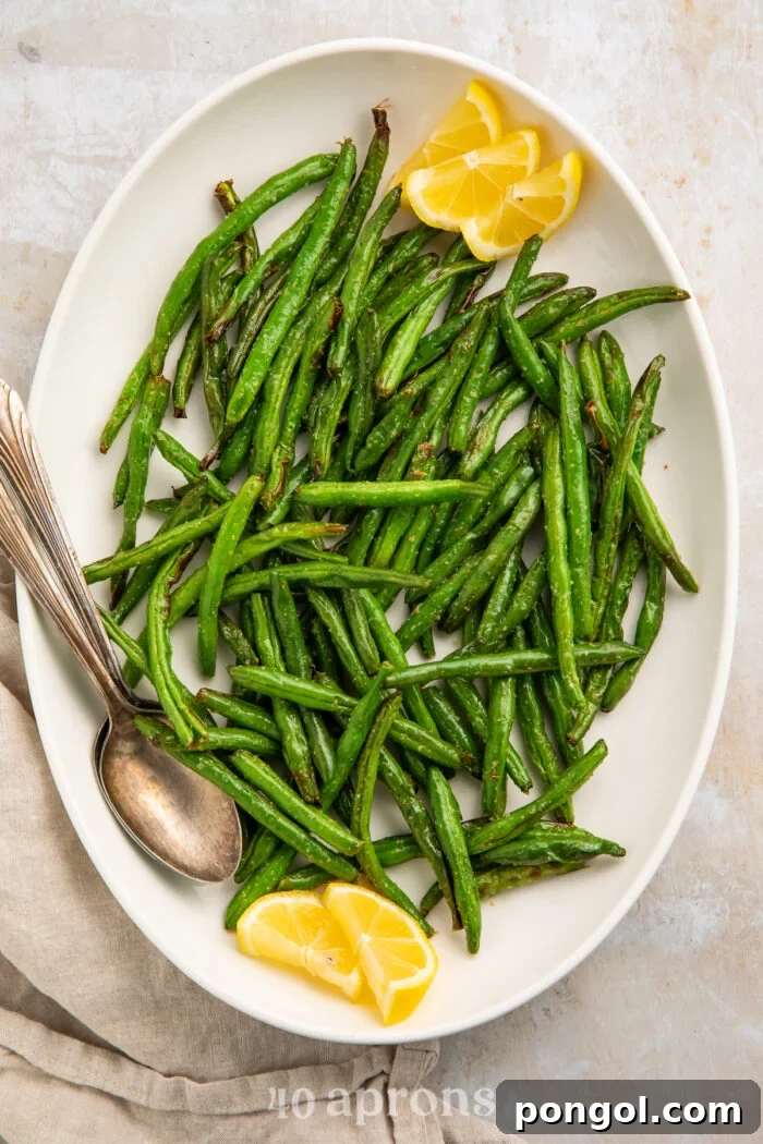Overhead view of bright air fryer green beans on a white platter with lemon wedges and a silver spoon.