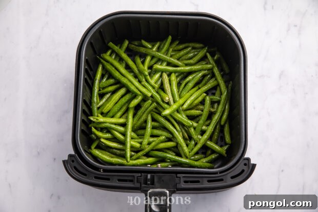 Overhead view of seasoned green beans in an air fryer basket on a white counter.