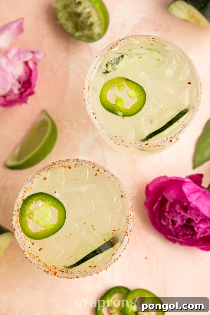 Jalapeño Cucumber Zing Margarita 3 Overhead view of two refreshing cucumber jalapeno margaritas on a rustic wooden table, artfully arranged with fresh cucumber slices and vibrant fuschia flowers.