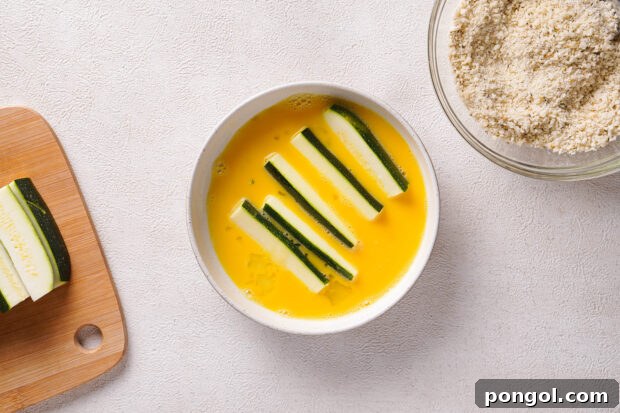 Overhead view of zucchini sticks in egg wash in small ceramic white bowl.