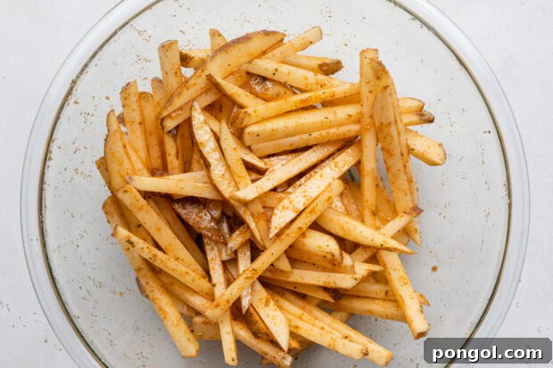 Overhead view of uncooked potato sticks tossed in cajun seasoning and avocado oil in a large glass mixing bowl.