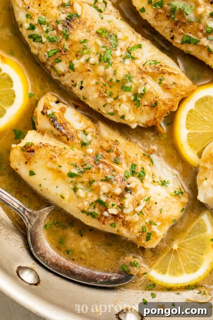 Overhead view of garlic butter cod fillets in a silver skillet with garlic butter sauce and lemon coins.