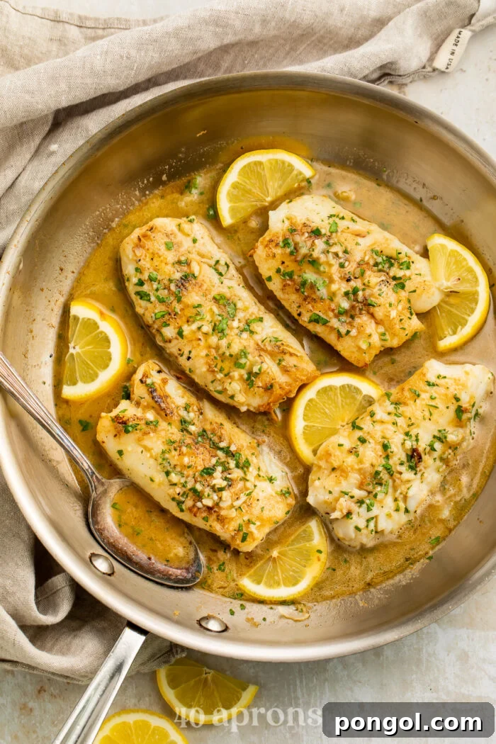 Overhead view of a skillet of cod fillets in a garlic butter sauce with lemon coins on a table.