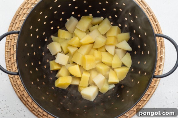 Overhead view of boiled potato cubes in a large black colander on a neutral background.