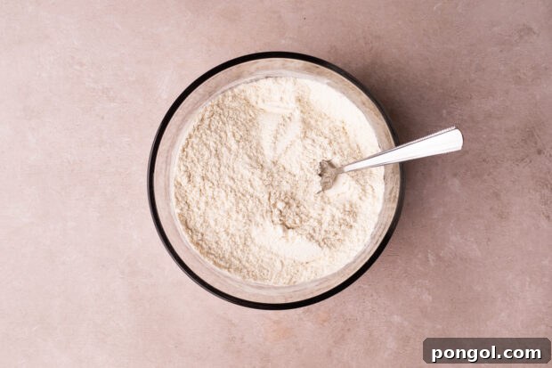 Effortless Air Fryer Chocolate Chip Cookies 6 Overhead view of flour, baking soda, and salt whisked together with a fork in a large glass mixing bowl on a pink surface.