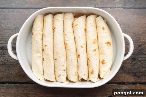 Overhead view of 7 rolled enchiladas neatly arranged in a white oval casserole dish on a neutral wooden background.