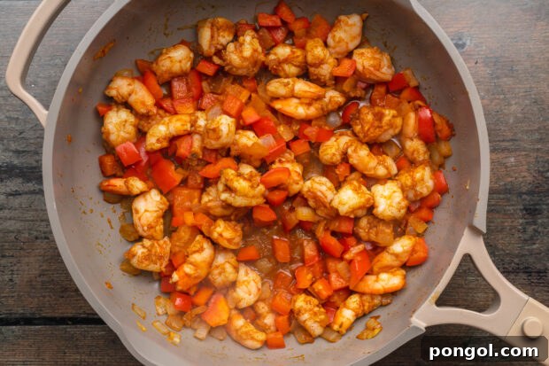Overhead view of chopped shrimp cooking in a large skillet with diced onion and red bell pepper.