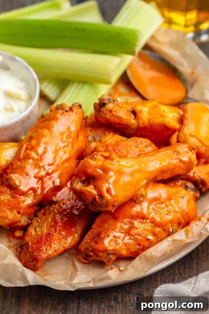 Overhead view of chicken wings coated in a glistening orange buffalo sauce on a sheet of tan parchment paper with bright green celery in the background.