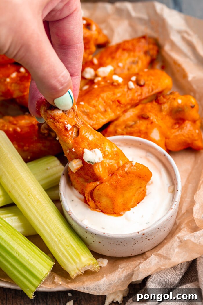 A white woman's hand dips an orange chicken wing in a small bowl of white ranch dressing that sits next to sticks of green celery.