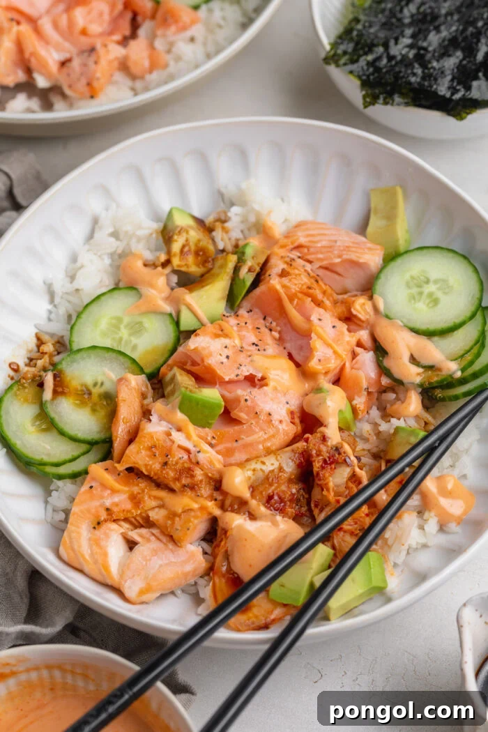 Angled view of a salmon rice bowl with salmon, rice, cucumber, avocado, seaweed, and chopsticks on a grey table.